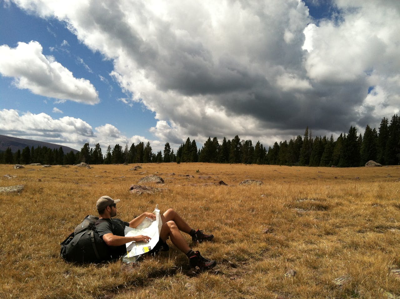 digital A man with a backpack reads a map in an open field with cloudy skies and trees in Daggett County, Utah.