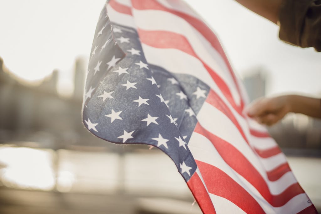 photo by ketut subiyanto From below of crop person holding national flag of United States of America waving in wind on street against city river